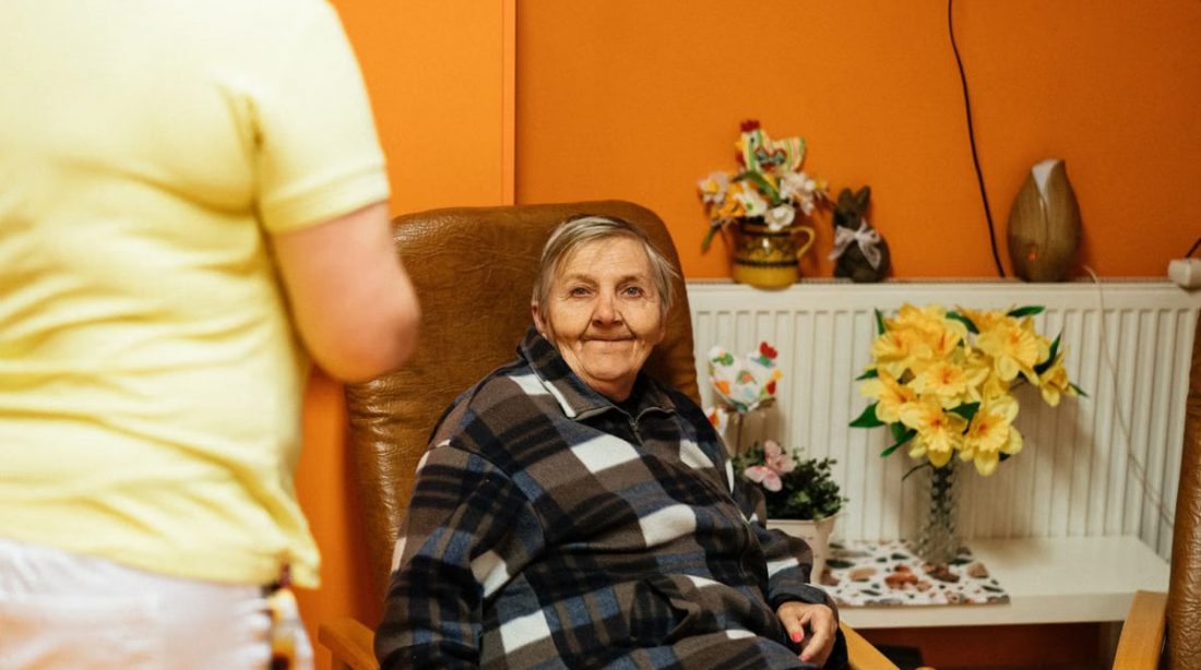 A warm scene of a senior woman sitting in an armchair at a retirement home with a caretaker nearby.