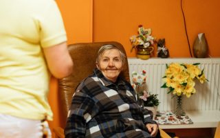 A warm scene of a senior woman sitting in an armchair at a retirement home with a caretaker nearby.