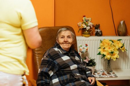 A warm scene of a senior woman sitting in an armchair at a retirement home with a caretaker nearby.