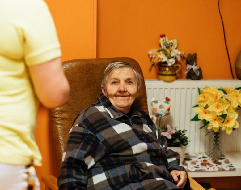 A warm scene of a senior woman sitting in an armchair at a retirement home with a caretaker nearby.